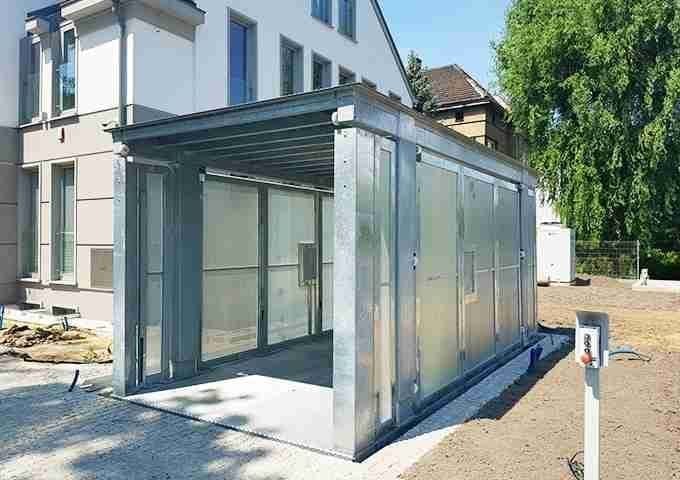 Estructura metálica moderna con puertas correderas de vidrio y ventanas conectada a una casa residencial blanca, jardín con césped y árboles de fondo