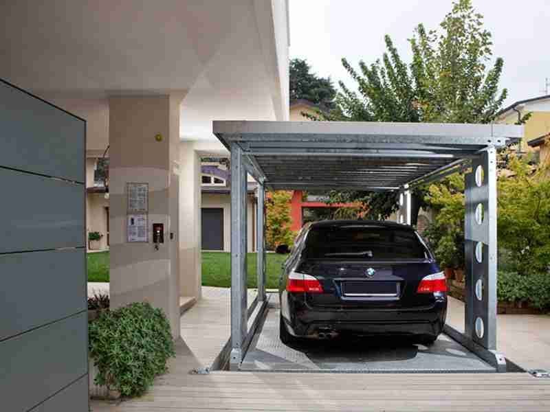 Coche negro aparcado bajo estructura metálica gris con techo, jardín y casa al fondo